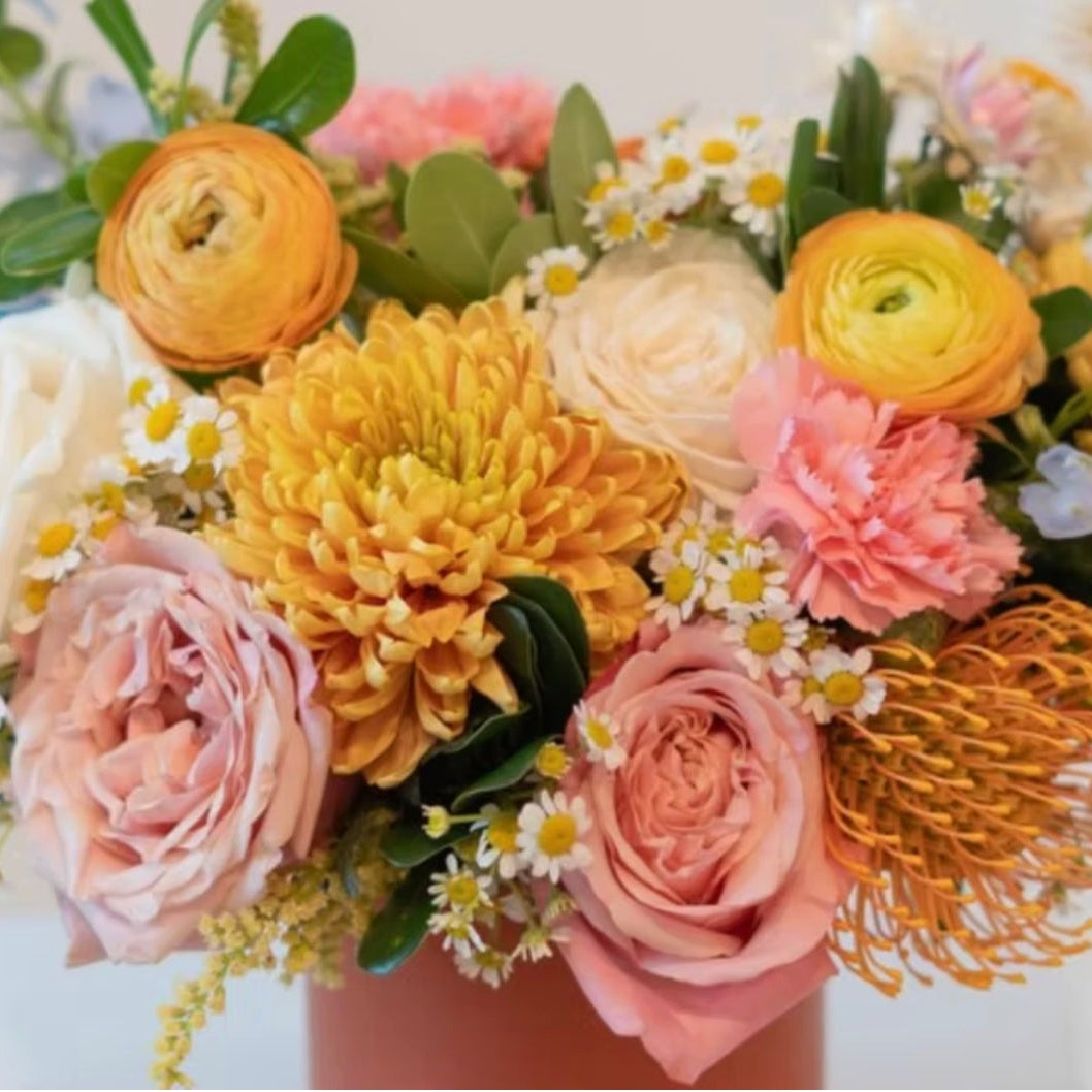 A colorful floral arrangement featuring pink, yellow, and white flowers with green foliage, presented in a soft pink container against a light background.